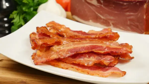 Knusprig gebratener Bacon im Ofen, auf einem weißen Teller gestapelt. Im Hintergrund ein Stück rohes Fleisch, Knoblauch und Petersilie.