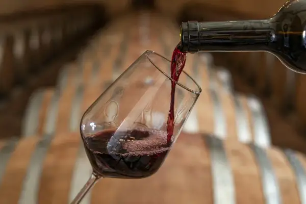 Versando vino rosso in un bicchiere, con botti di legno sullo sfondo. Attenzione, il vino contiene nichel.
