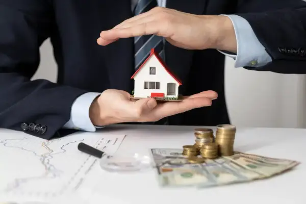 Mani che proteggono una casa modello, con grafici, soldi e monete. L'idea di comprare casa per affittarla è ben rappresentata.