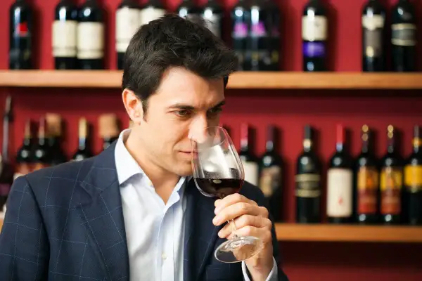 Mężczyzna w garniturze wącha czerwone wino w kieliszku. To degustator wina, który ocenia jego aromat.