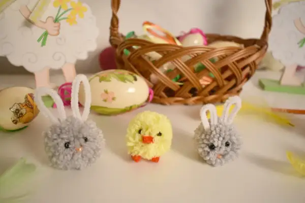 Urocze ozdoby wielkanocne zrób to sam dla dzieci: puchate króliczki i kurczaczek z pomponów, obok pisanki i koszyczek.