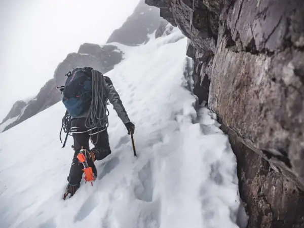 Grimpeur en crampons et piolet, progressant sur une pente enneigée. Un entraînement en altitude exigeant, au cœur d'un paysage rocheux et brumeux.