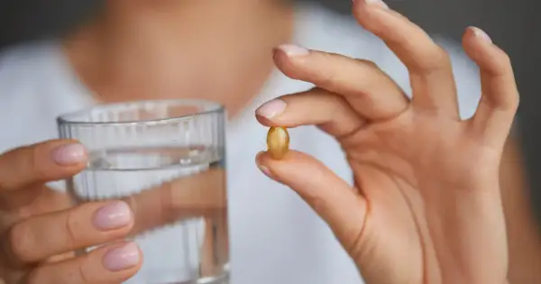 Dłoń trzyma kapsułkę omega 3 i szklankę wody. Kiedy brać omega 3? Najlepiej z posiłkiem.