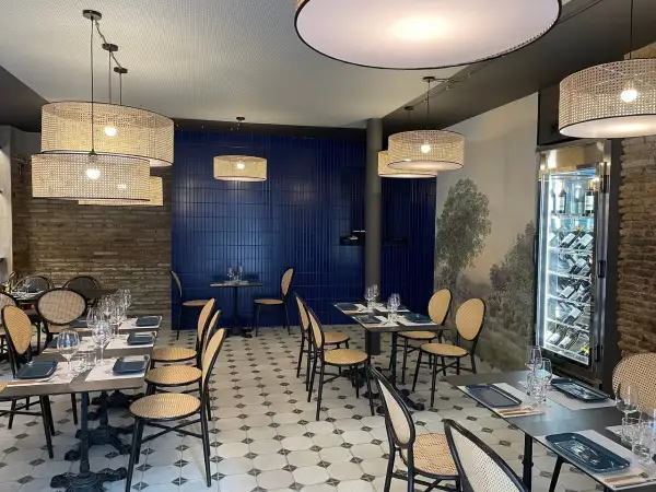 Interior de la brasserie 1885, con mesas puestas, sillas de mimbre y lámparas de ratán. Pared azul y mural de paisaje.