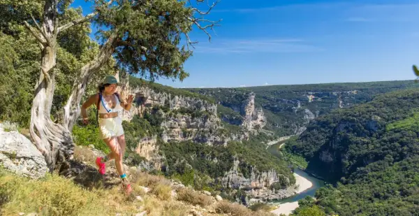 Une femme court sur un sentier escarpé avec une vue spectaculaire sur le **trail gorge de l'Ardèche**. Le soleil brille sur les falaises et la rivière.
