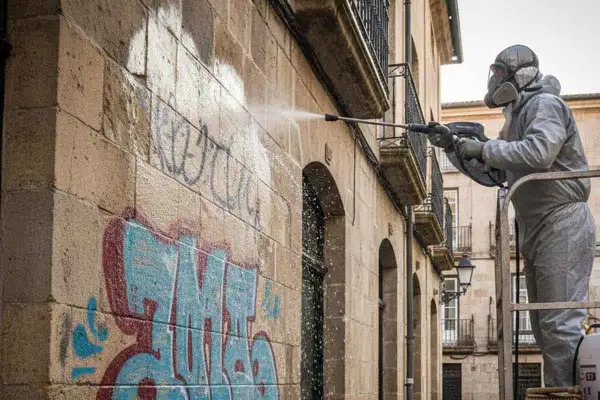 Trabajador con traje protector realiza limpieza de fachada con hidrolimpiadora, eliminando grafitis de un muro de piedra.