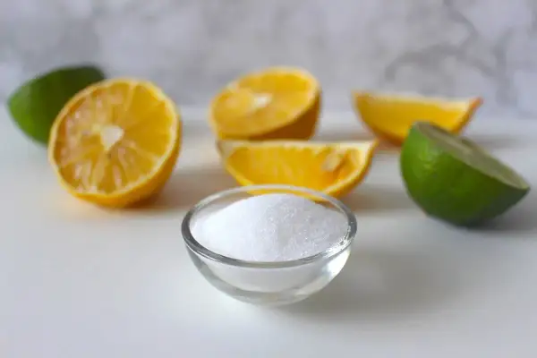 Ciotolina di acido citrico cristallino circondata da spicchi di limone e lime, ingredienti freschi per un tocco frizzante.