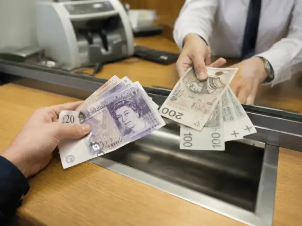 Ręka podaje wycofane funty i polskie banknoty w okienku bankowym.