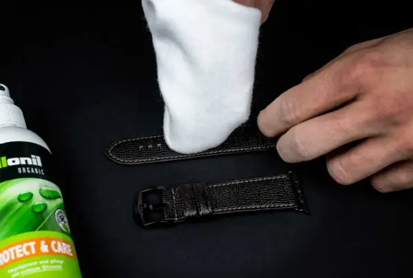 Ein weißes Tuch poliert ein dunkles Lederarmband. Daneben steht eine Flasche "Collonil Organic Protect & Care" zur Pflege.