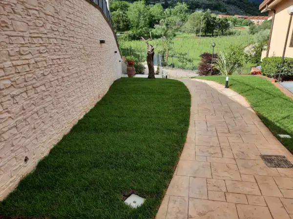 Sentiero in cemento stampato spessore minimo, con finitura effetto pietra, fiancheggiato da prato verde e muro in pietra grezza.