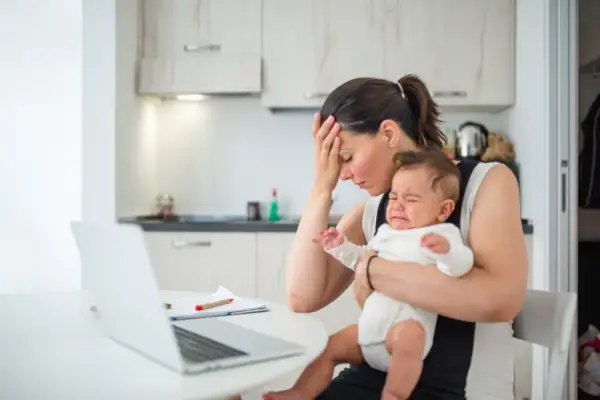 Zmęczona mama próbuje pracować przy laptopie, trzymając płaczącego, nadpobudliwego niemowlaka.