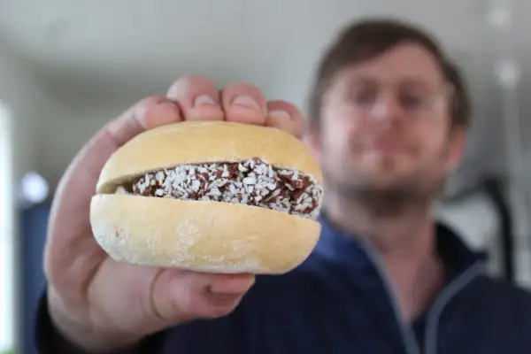 En hand håller upp en bulle med fyllning av choklad och kokosflingor. En man syns i bakgrunden. En riktig bulle med bulle!