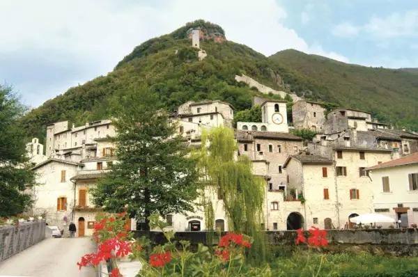 Antico borgo arroccato su un colle verdeggiante, uno dei paesi della Valnerina, con case in pietra, un campanile con orologio e una torre medievale in cima.