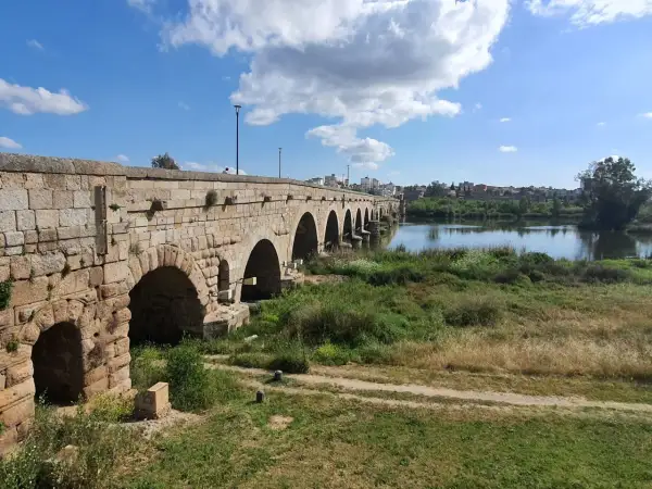 Puente romano de piedra con arcos sobre un río, bordeado de vegetación. Un camino de tierra serpentea por la orilla, evocando la antigua **Vía de la Plata**.