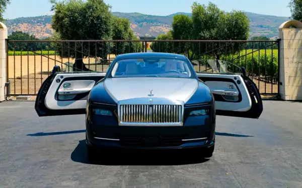 Elegancki Rolls-Royce Spectre z otwartymi drzwiami, zaparkowany przed bramą posiadłości z widokiem na wzgórza.