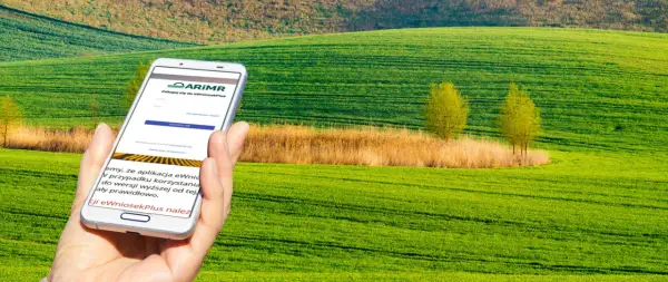 Dłoń trzyma smartfon z aplikacją ARiMR do składania wniosków o płatności bezpośrednie na tle zielonych pól.