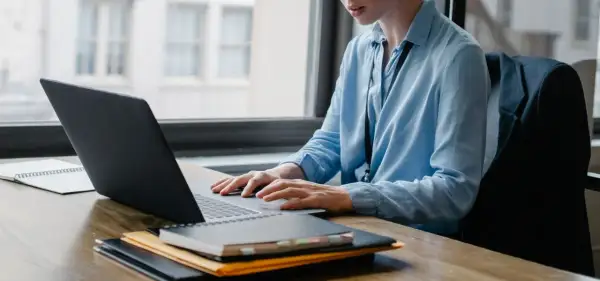 Osoba w niebieskiej koszuli pracuje przy laptopie, tworząc skrypty VBA. Na biurku leżą notatniki.