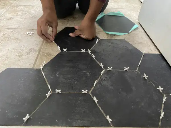 Hands place dark hexagonal tiles over existing floor tiles, using white spacers to ensure even gaps.