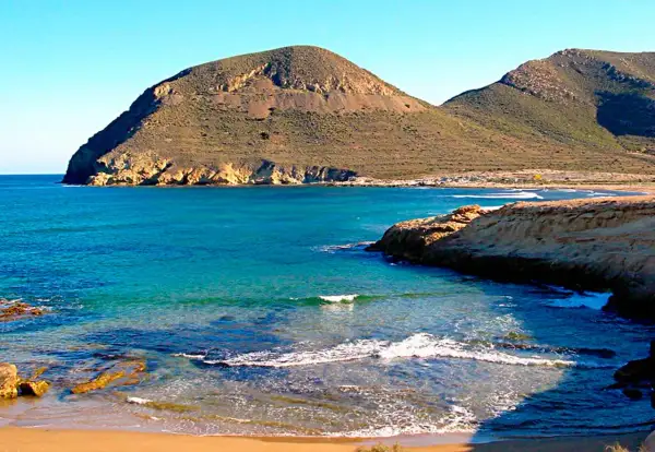 Aguas turquesas bañan la costa rocosa de el playazo las negras, con montañas áridas de fondo bajo un cielo azul.