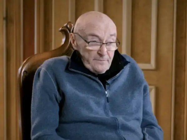 Phil Collins w okularach, siedzi w fotelu. Jego choroba wpływa na jego zdolność do gry na perkusji, ale nadal inspiruje fanów.