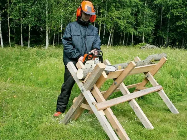 Man med motorsåg använder en hemmagjord sågbock för att kapa ved i en grönskande skogsmiljö.
