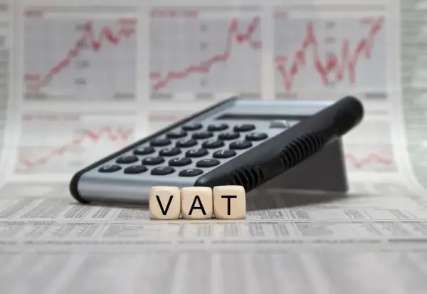 Kalkulator i kostki z literami VAT na tle wykresów finansowych. Pełna księgowość krok po kroku.