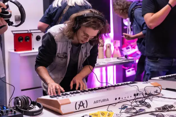 Młody mężczyzna w słuchawkach gra na keyboardzie Arturia. W tle widać sprzęt audio i innych ludzi, tworząc atmosferę pracy w wytwórni muzycznej.