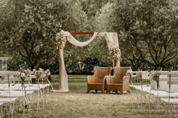 Un bel exemple de cérémonie laïque en plein air, avec un arche fleuri, des chaises blanches et deux fauteuils en rotin sous les oliviers.