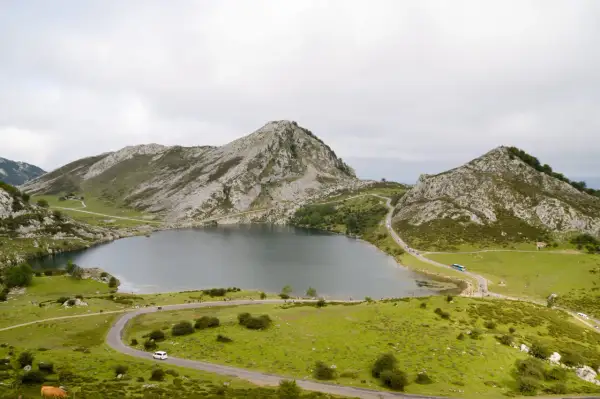 Paisaje montañoso con un lago sereno y carreteras sinuosas. Se puede ver gente disfrutando del **subir Lagos de Covadonga**.