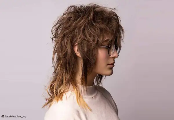 En ung kvinna med glasögon bär en shaggy mullet frisyr med rufsigt, brunt hår som bleknar till blont i topparna.
