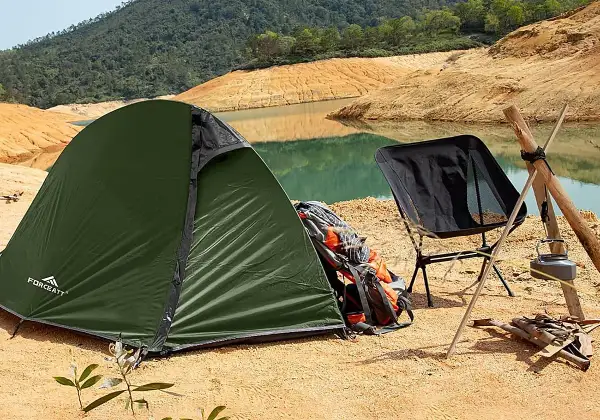 Tienda de campaña verde junto a un lago, con una silla plegable negra y mochilas de transporte.