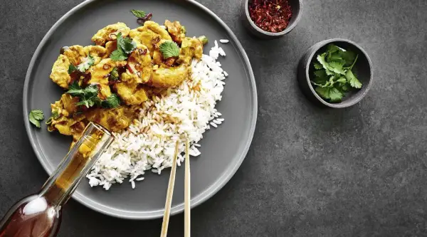 Ein Teller mit Reis und einem köstlichen Thai Curry, garniert mit frischem Koriander. Daneben kleine Schälchen mit Chiliflocken und Koriander.