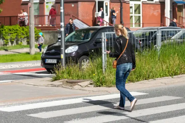 Młoda kobieta, pieszy, idzie przez pasy, patrząc w telefon.