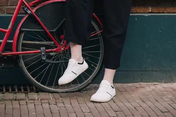 Voeten in witte barefoot schoenen naast een rode fiets. Nadelen barefoot schoenen: minder bescherming op oneffen ondergrond.