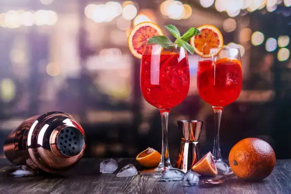 Dwa czerwone drinki z pomarańczą i miętą, idealne do prosecco. Obok shaker i pomarańcze.