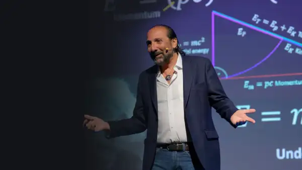 Nassim Haramein, con gestos amplios, explica conceptos de física ante una pantalla con fórmulas como E=mc².