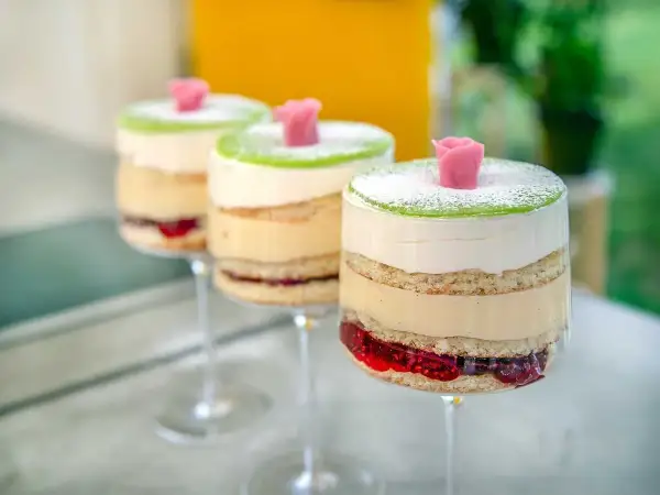Tre vackra prinsesstårtor i glas, med lager av sockerkaka, grädde, vaniljkräm och hallonsylt, toppade med grön marsipan och en rosa ros.