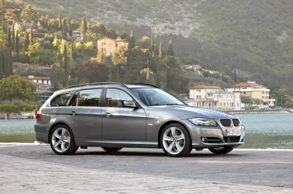 Szary kombi BMW 325i zaparkowany nad jeziorem, z malowniczymi wzgórzami i budynkami w tle.