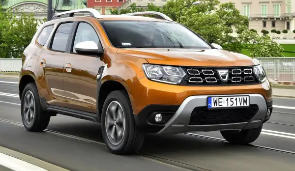 Pomarańczowy Dacia Duster 2 jedzie po moście, za nim widać budynki i drzewa.