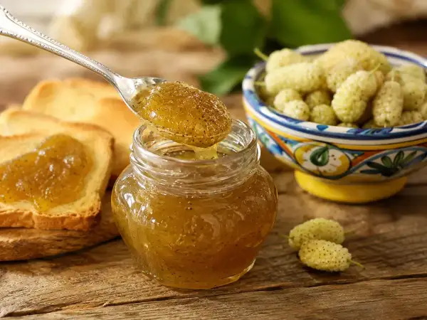Marmellata di gelsi bianchi su pane tostato, con una ciotola di frutti freschi e un cucchiaino che ne raccoglie una porzione.