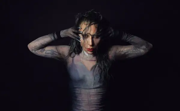 Lady Gaga w siateczkowej bluzce z długimi rękawami i tatuażami, zakrywająca twarz dłońmi.