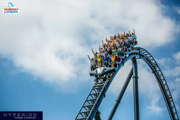 Ludzie krzyczą z radości na pokładzie Hyperiona, największego rollercoastera w Polsce, pędząc po stalowych torach na tle błękitnego nieba.