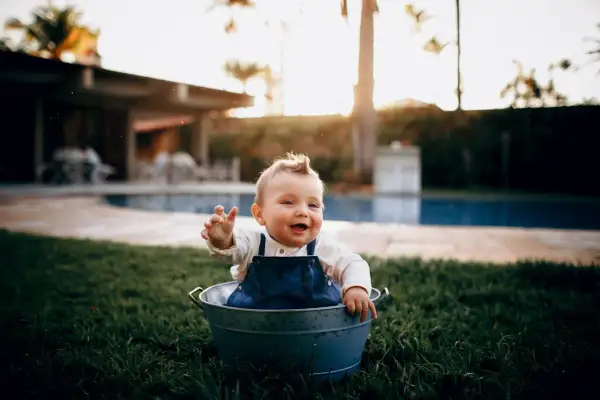 Glad bebis i en hink på gräsmattan, med en pool i bakgrunden. En härlig dag för ett bebis bada i poolen!