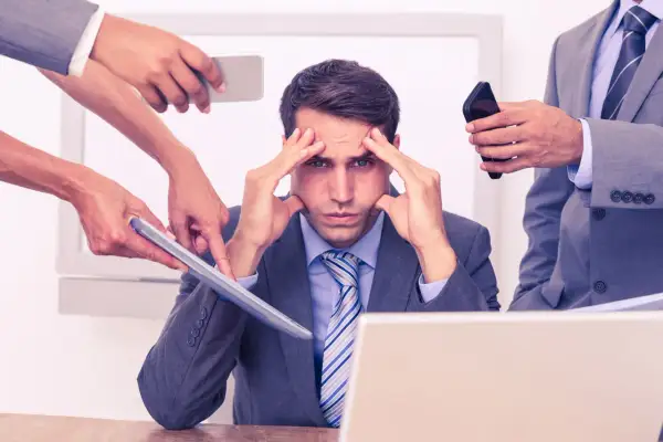 Stressad man med händer på tinningarna, omgiven av kollegor som ger instruktioner. Dålig kognitiv ergonomi leder till överbelastning.