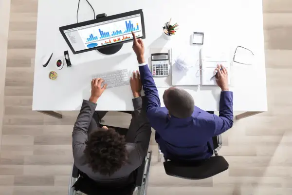 Två personer vid ett skrivbord analyserar data på en skärm, ett exempel på it-system. En person i rullstol skriver på tangentbordet, den andra pekar på diagrammet.