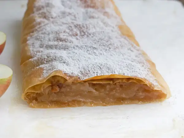 Ein köstlicher Apfelstrudel mit fertigem Strudelteig, bestäubt mit Puderzucker. Daneben liegen frische Apfelstücke.