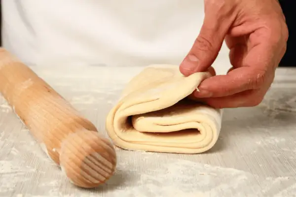 Mani che piegano la pasta sfoglia fatta in casa, con un mattarello in legno accanto, su un piano infarinato.