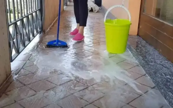 Pulizia del terrazzo esterno: una scopa blu lava le piastrelle con acqua saponata, accanto un secchio giallo.