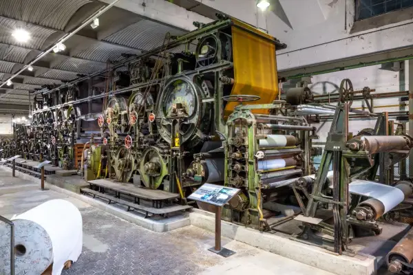 Große, grüne Textilmaschine im lvr industriemuseum, die Stoffbahnen verarbeitet. Eine gelbe Stoffbahn hängt oben, daneben Papierrollen.