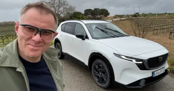 Mężczyzna w okularach pozuje do selfie obok białej Mazdy CX-5 na tle winnicy.